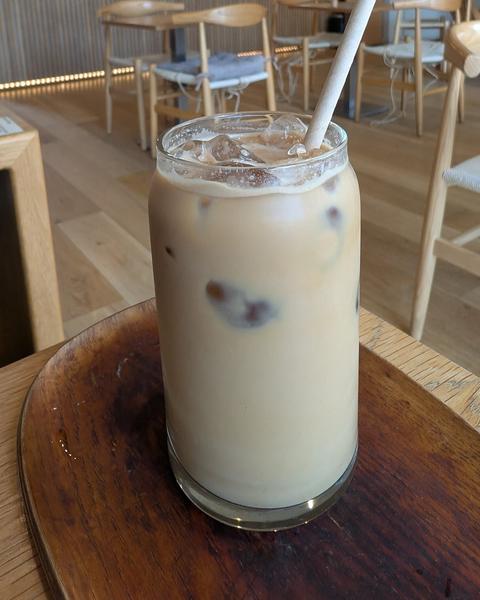 A tall glass of iced milky coffee with a paper straw, on a wooden board in a café