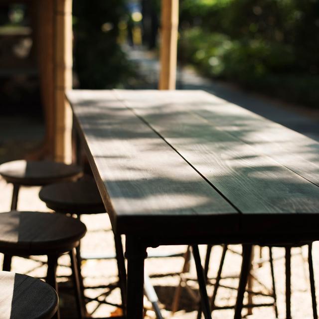 A long wooden table with black stools on a patio in dappled sunlight