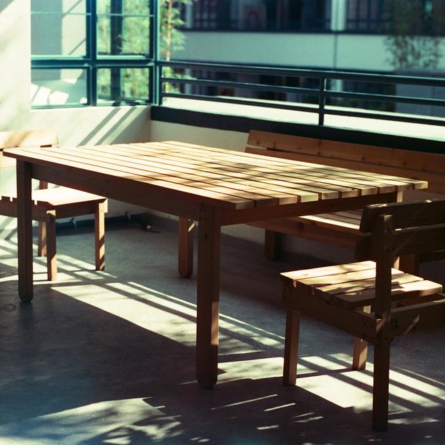 A wooden table and benches beside tall windows, with sunlight and shadows across the floor