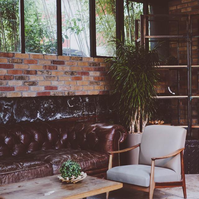 A corner with a leather bench, a pale armchair, a low wooden table and a plant against a brick wall