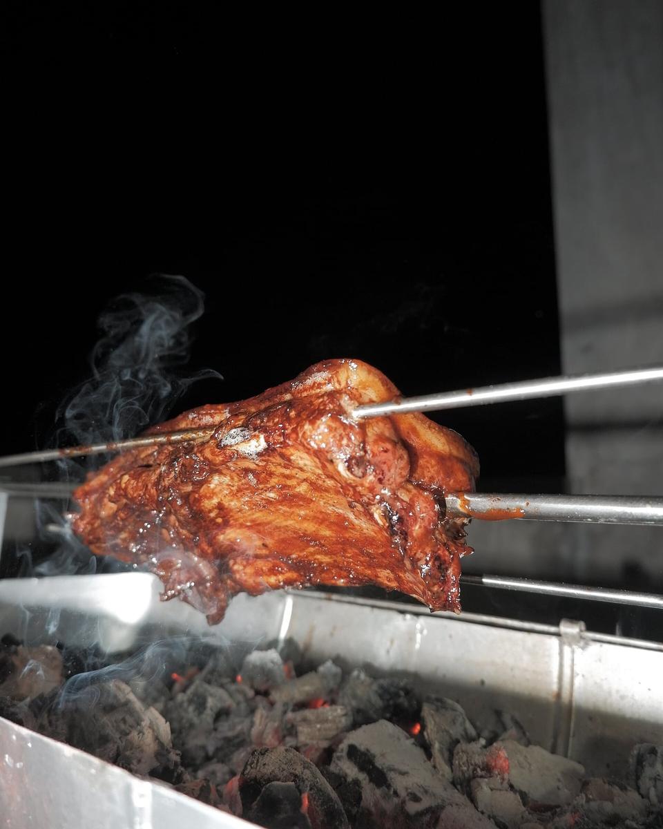 A slab of pork belly roasting on a spit over glowing charcoal, with smoke rising
