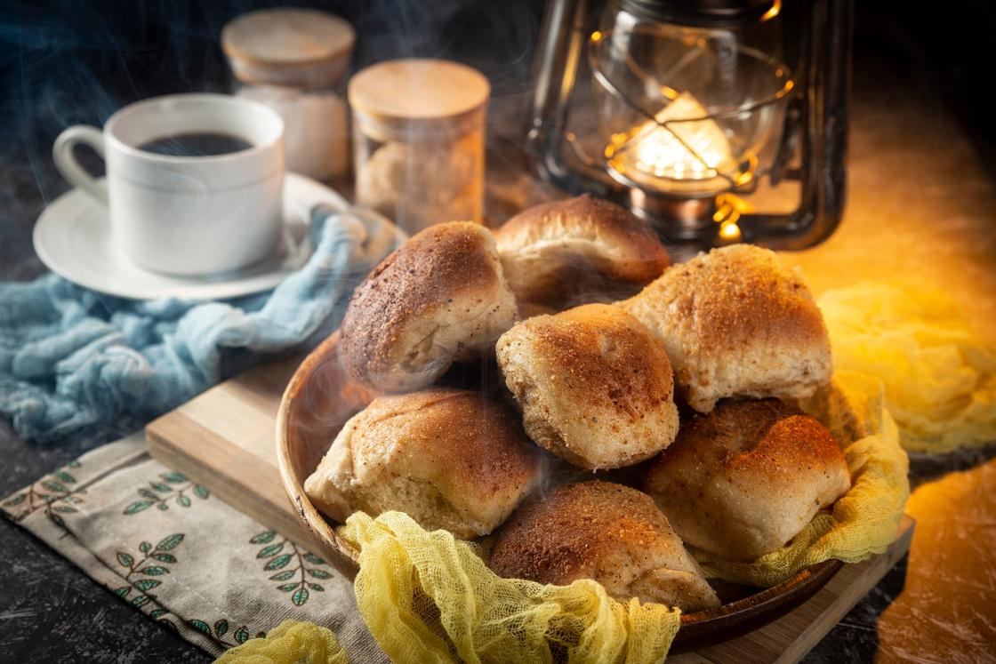A white cup of black coffee beside a basket of pandesal, with a lantern glowing behind